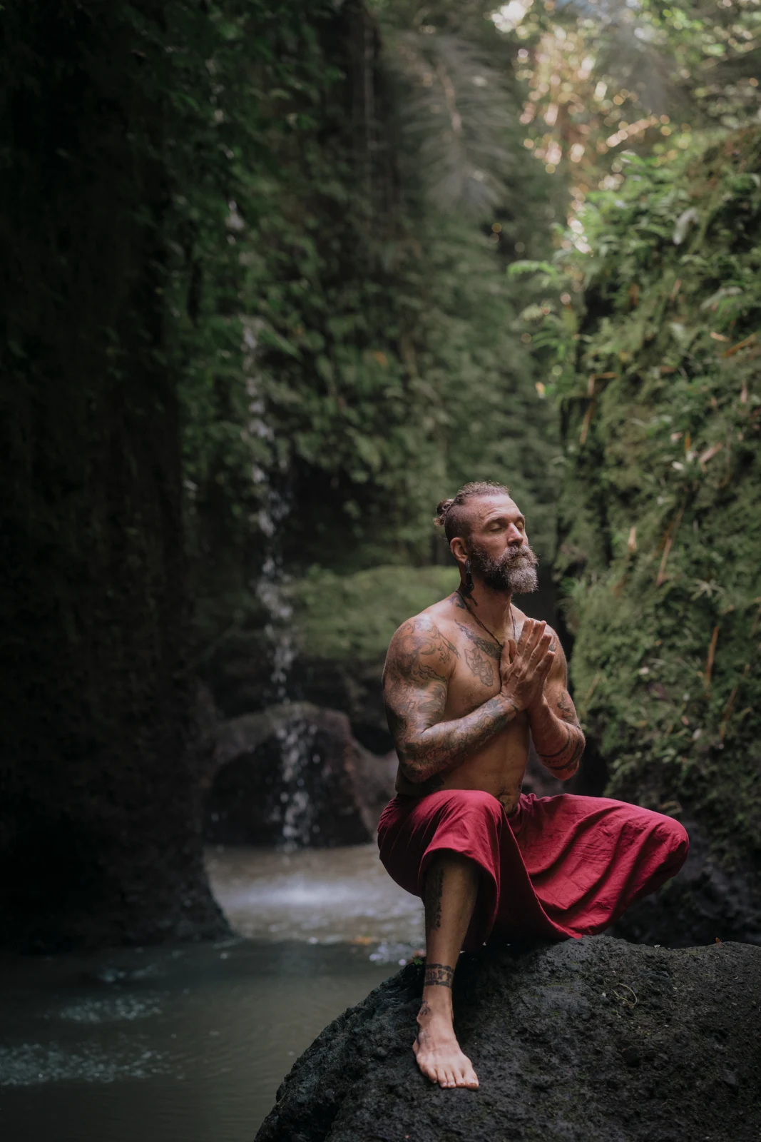 Ashton seated with hands together beside a waterfall in a green gorge.