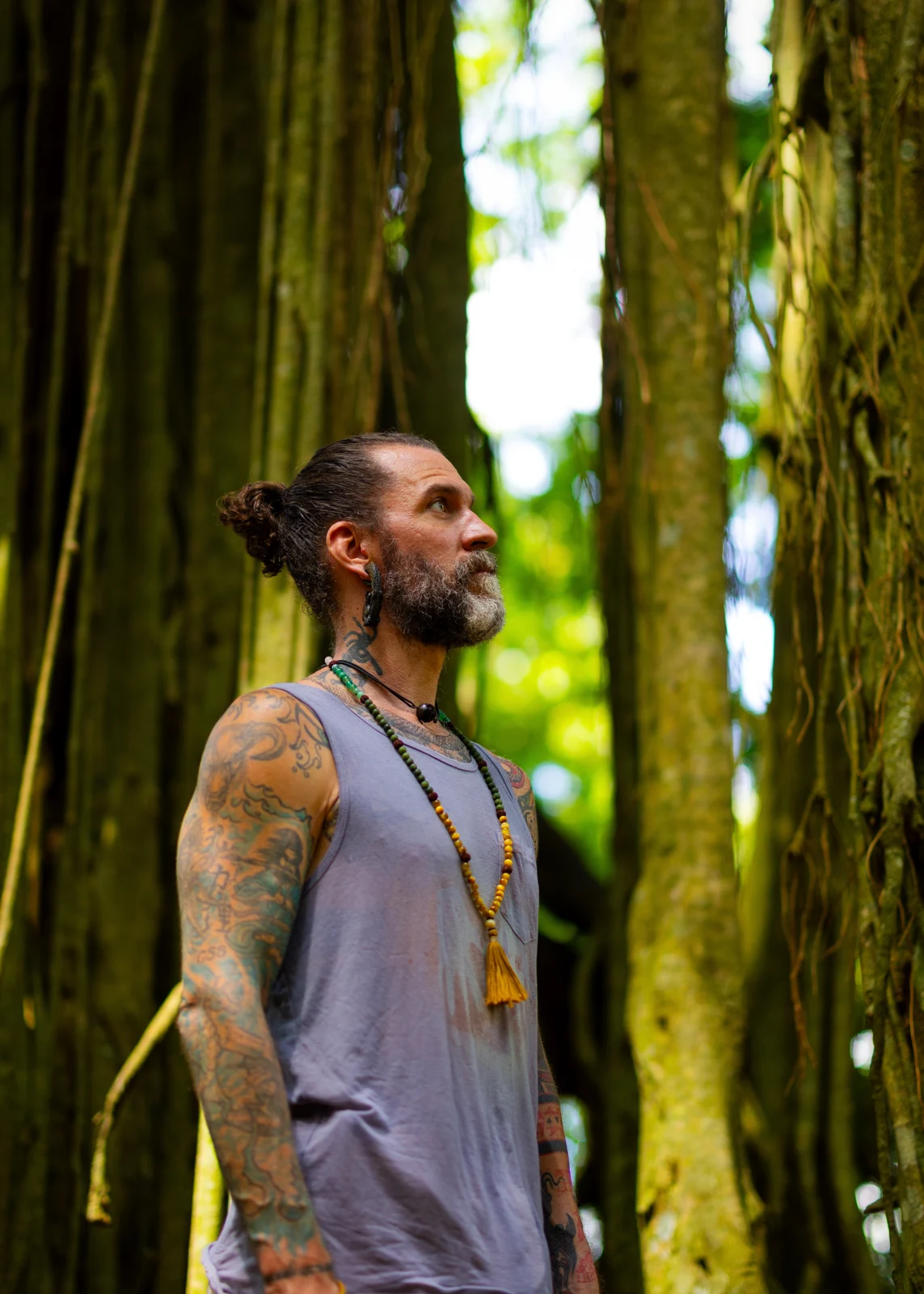 Ashton wearing a mala, looking upward among tree roots.